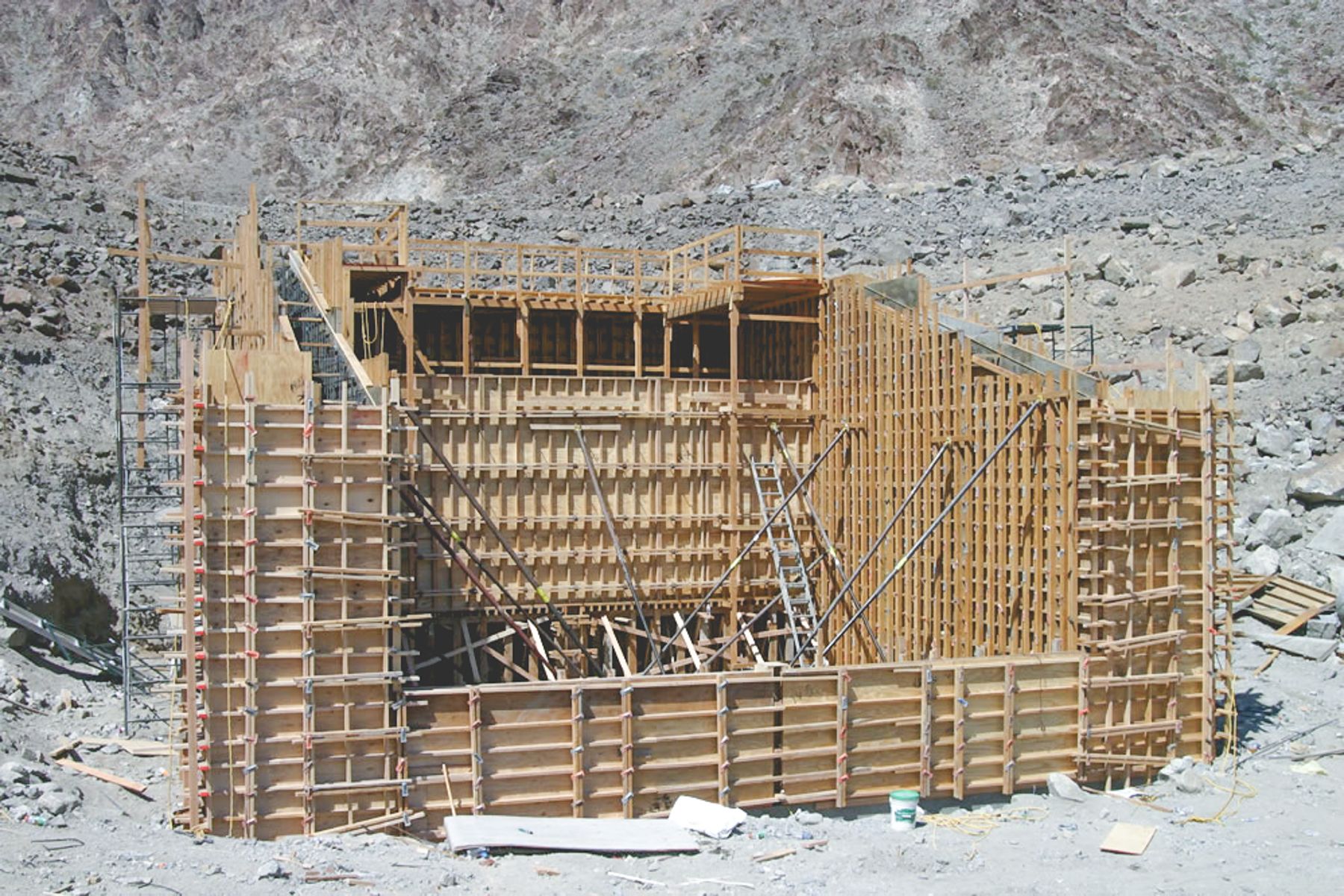 Tall concrete structure enclosed in plywood formwork with walers, tie rods, diagonal braces and a scaffold stair on a rocky slope