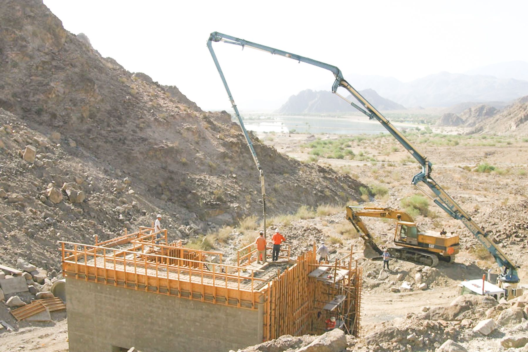 Truck-mounted concrete pump boom reaching over a formed concrete structure on a rocky slope while a crew places concrete on the top deck