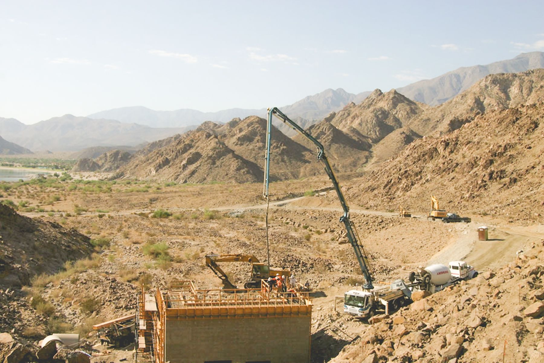 Boom pump placing concrete into a formed structure on a desert hillside job site, with an excavator and crew working alongside
