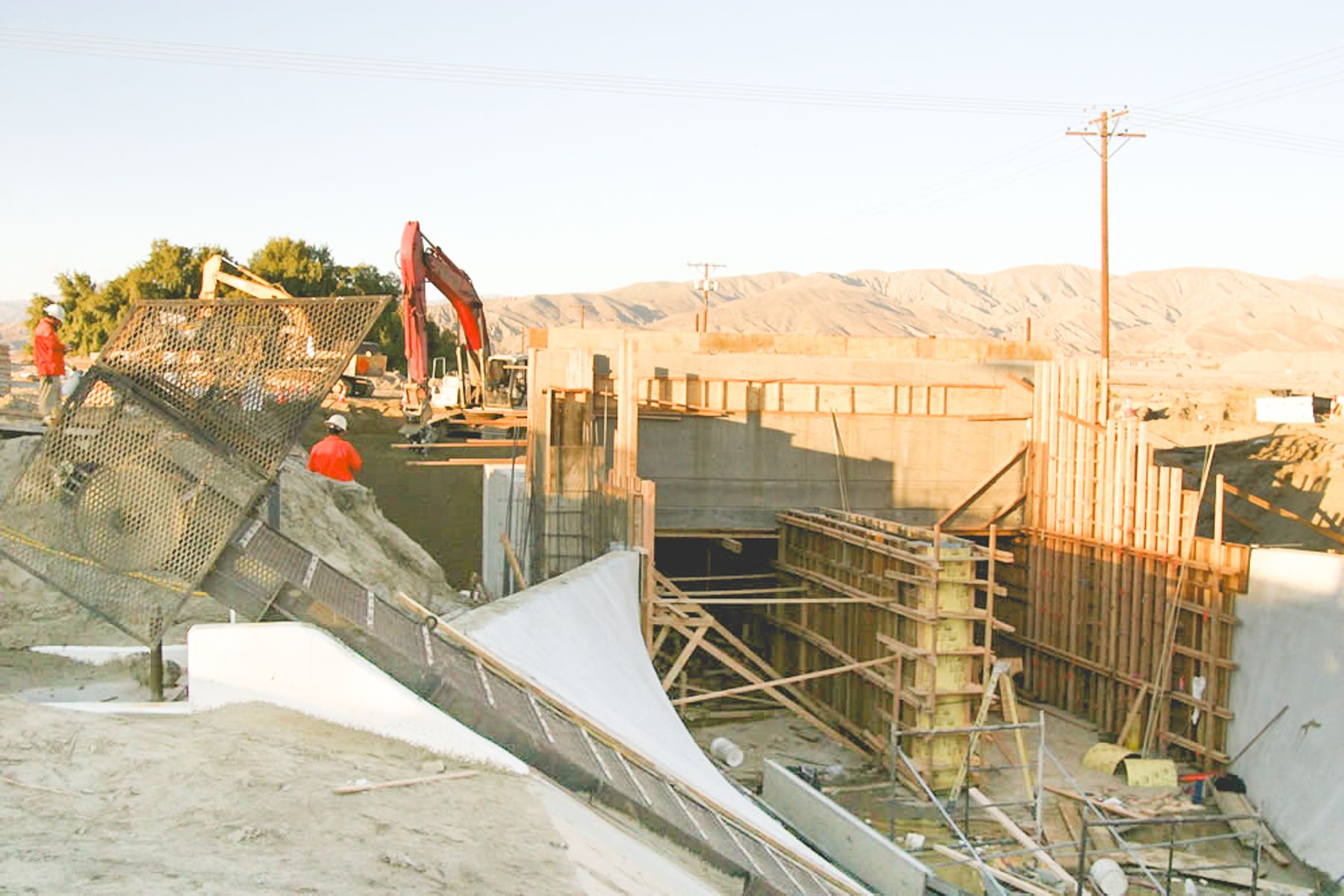 Formed concrete walls and a sloped wing wall at the canal crossing, with a red excavator and crew working at the top of the excavation