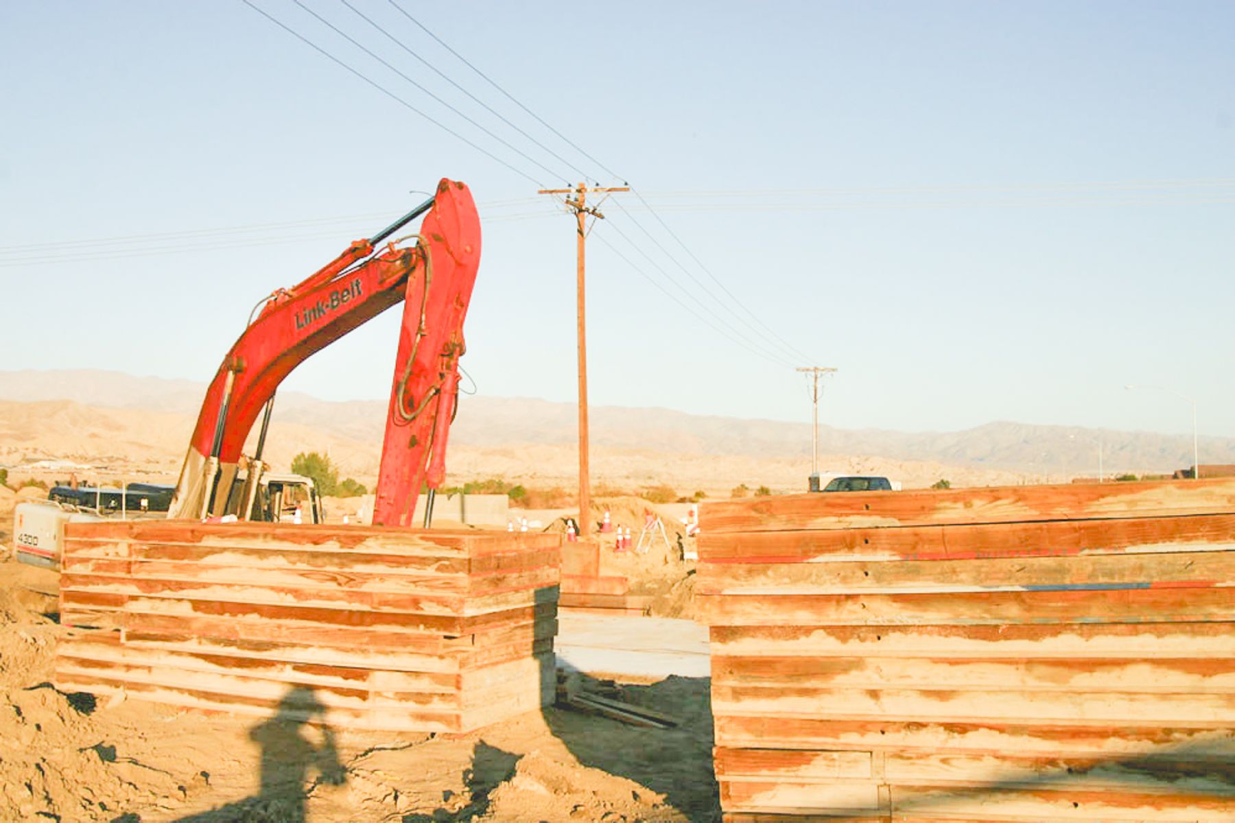 Stacked timber shoring panels in front of a red Link-Belt excavator, with utility poles and desert hills behind