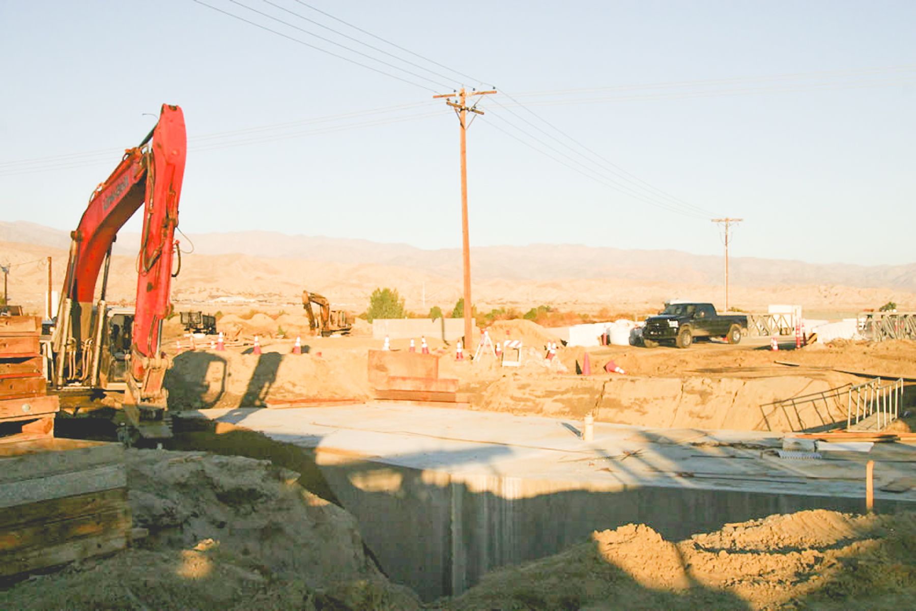 Concrete structure in an open excavation, with a red excavator, a pickup truck and forms at the top of the cut