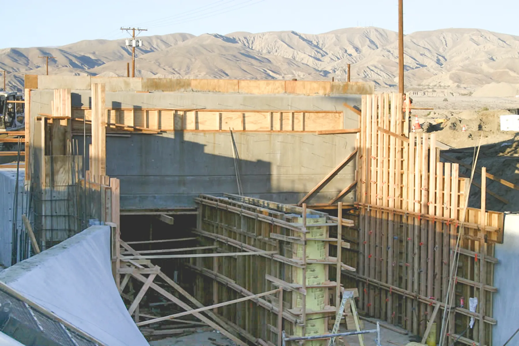 Cast-in-place concrete structure mid-construction, with timber formwork and bracing in the openings, white-coated wing walls in front and desert mountains behind