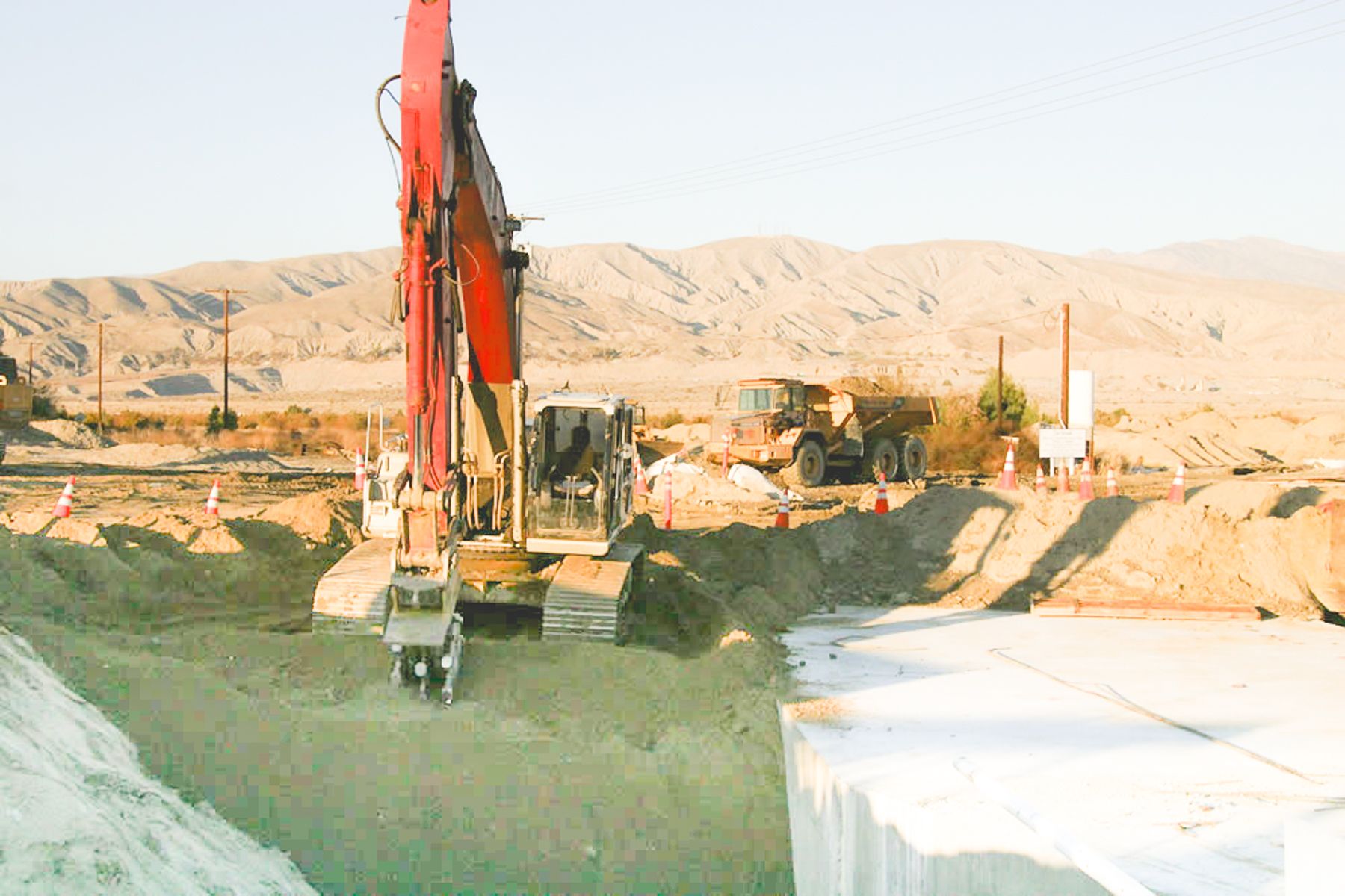 Red excavator working in the cut next to a white-coated concrete surface, with an articulated dump truck and cones behind
