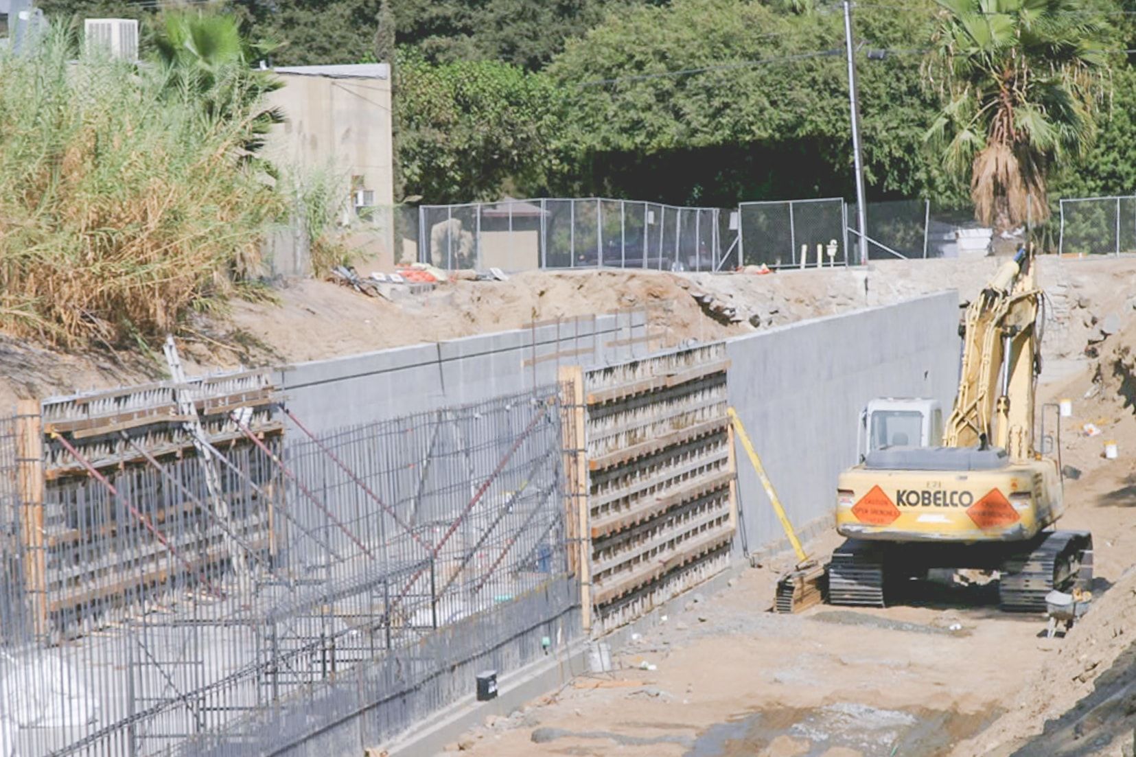 Concrete U-channel under construction beside a chain-link fence, with a rebar cage and wall forms on the left, a finished wall on the right and a Kobelco excavator on the channel floor