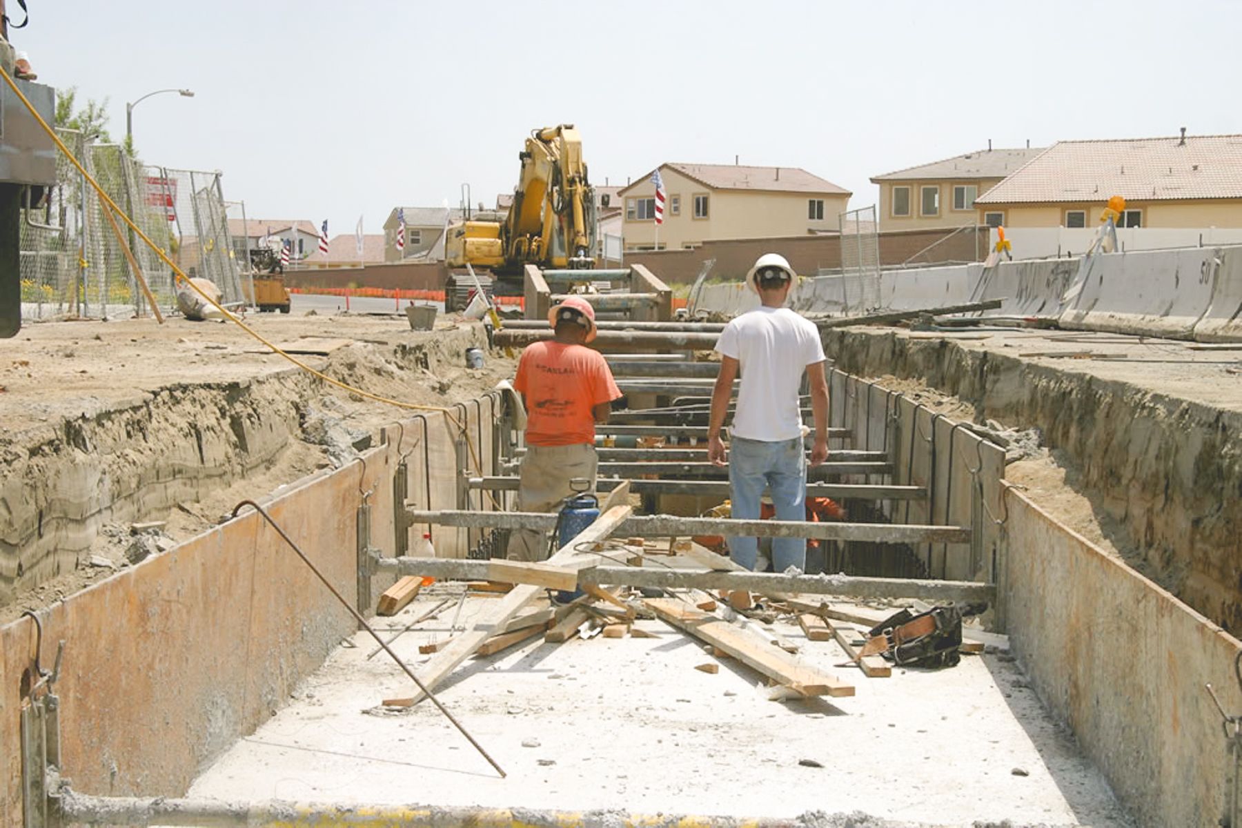 Two workers in hard hats standing on the poured base slab inside the shored trench, with timber and tools around them
