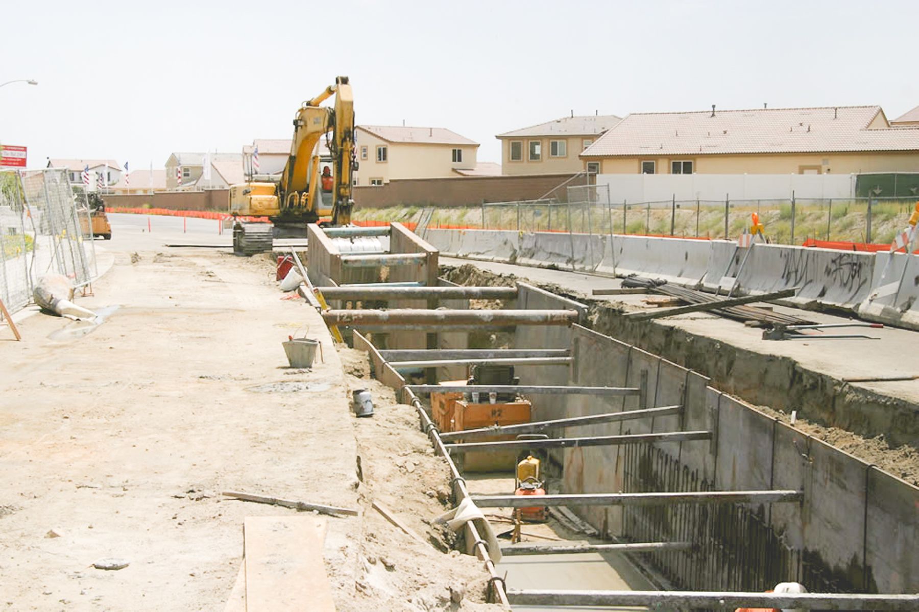 Long steel-shored trench with pipe cross-struts beside a new residential tract, with a yellow excavator at the far end