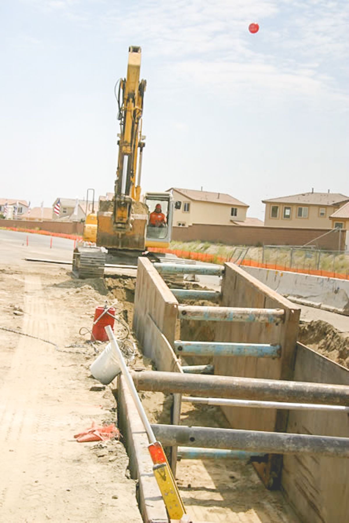 Yellow excavator at the end of the trench, with steel shoring panels and struts in the foreground and homes behind