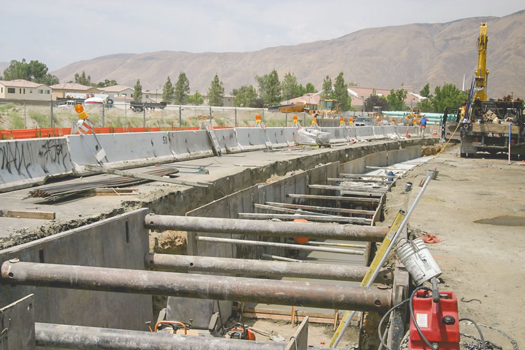 Curving shored trench running alongside K-rail and fencing, with steel plates, cross-struts and a work truck beyond