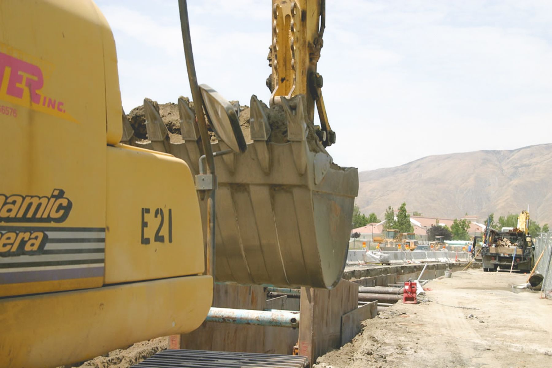 Close view of a yellow excavator marked E21 holding a loaded bucket over the shored trench, with hills behind