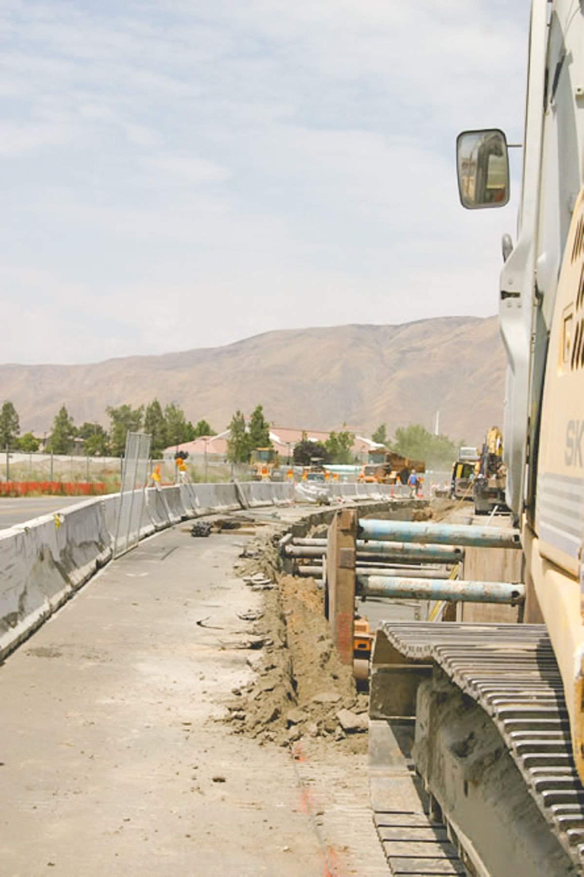 Trench shoring running along K-rail beside a paved road, seen past the tracks of an excavator