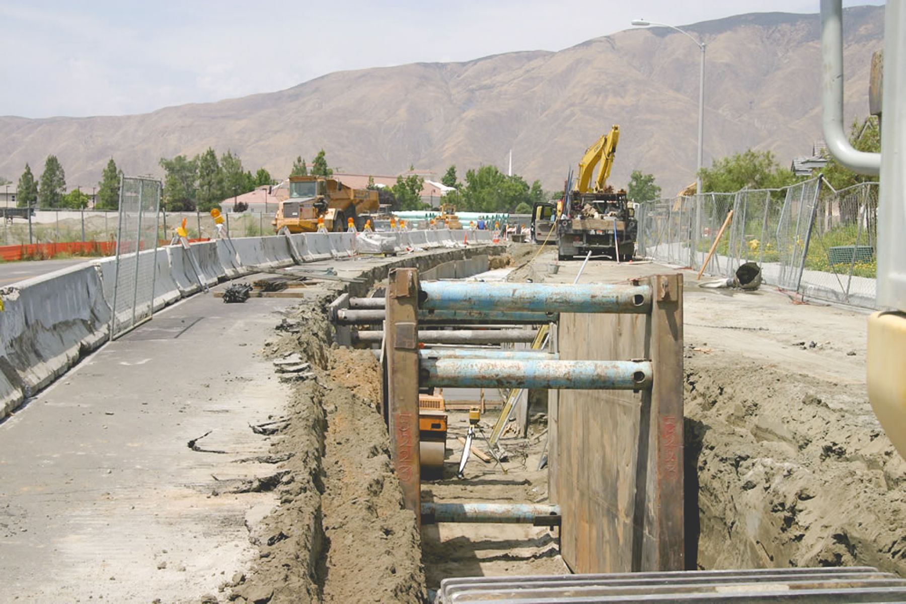 Close view of steel trench shoring with blue pipe struts beside a paved road, with an articulated dump truck and excavator in the distance
