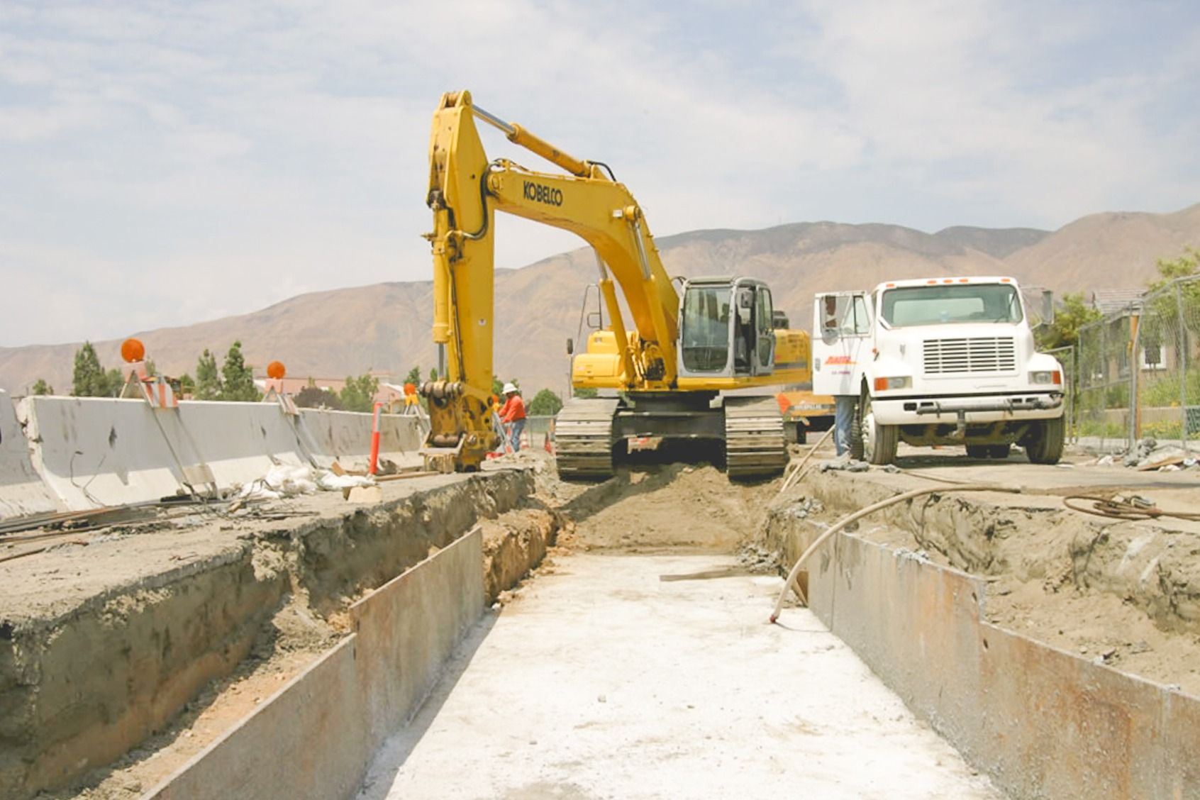 Yellow Kobelco excavator and a white work truck at the edge of a trench with a white-coated concrete base slab