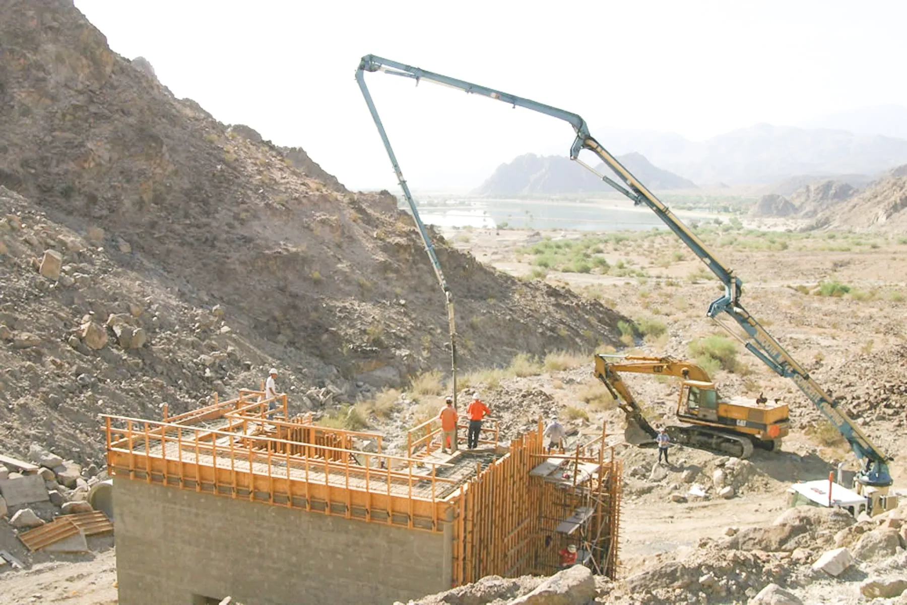 Concrete boom pump placing the top deck of a formed hillside dissipator structure while an excavator waits nearby in Coachella